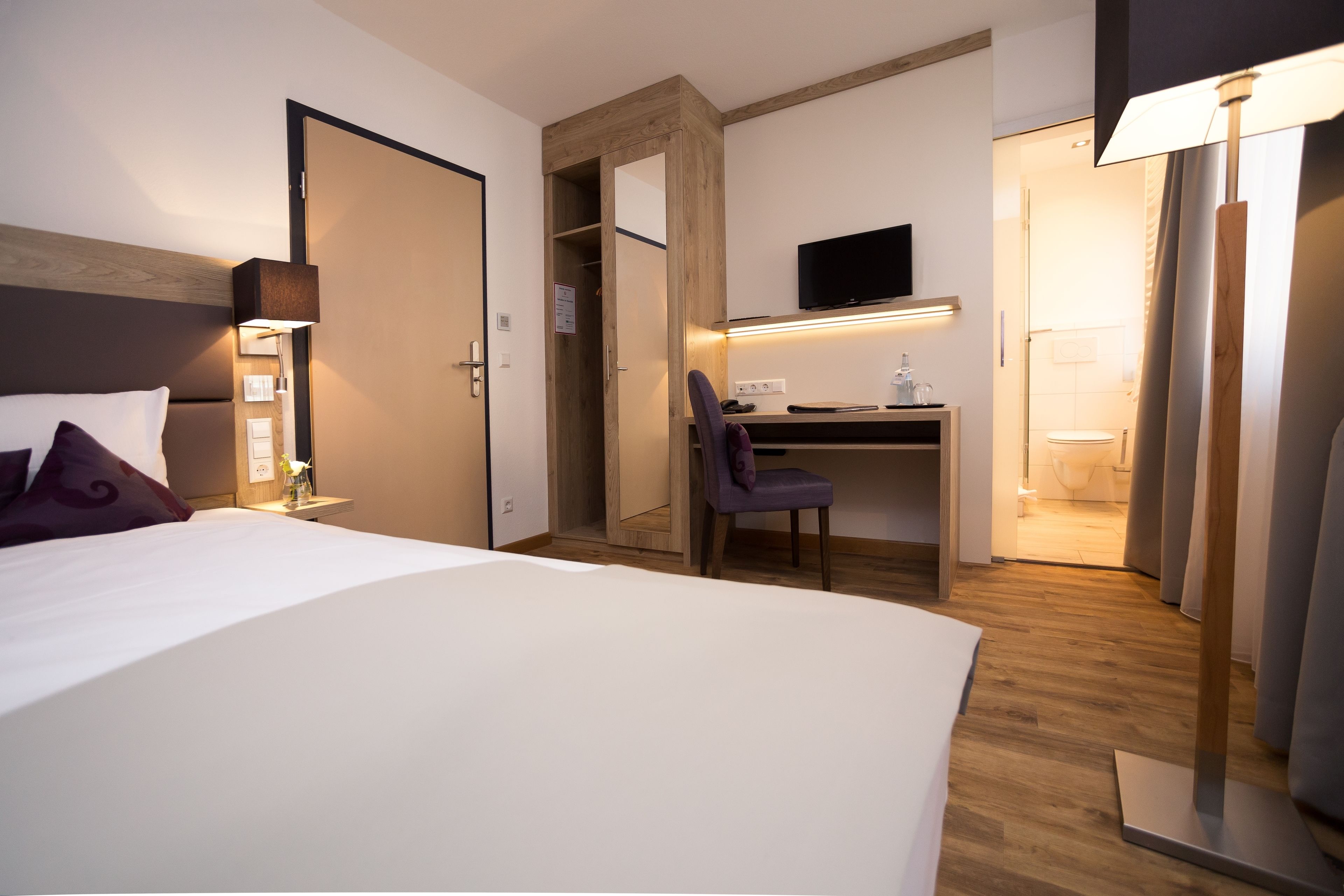 A hotel room with a bed, a desk with a chair, a TV mounted on the wall, and a bathroom visible through an open door.