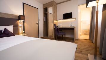A hotel room with a bed, a desk with a chair, a TV mounted on the wall, and a bathroom visible through an open door.
