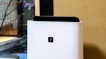 A white air purifier with the brand name "SHARP" on its base, placed on a textured surface near a window.
