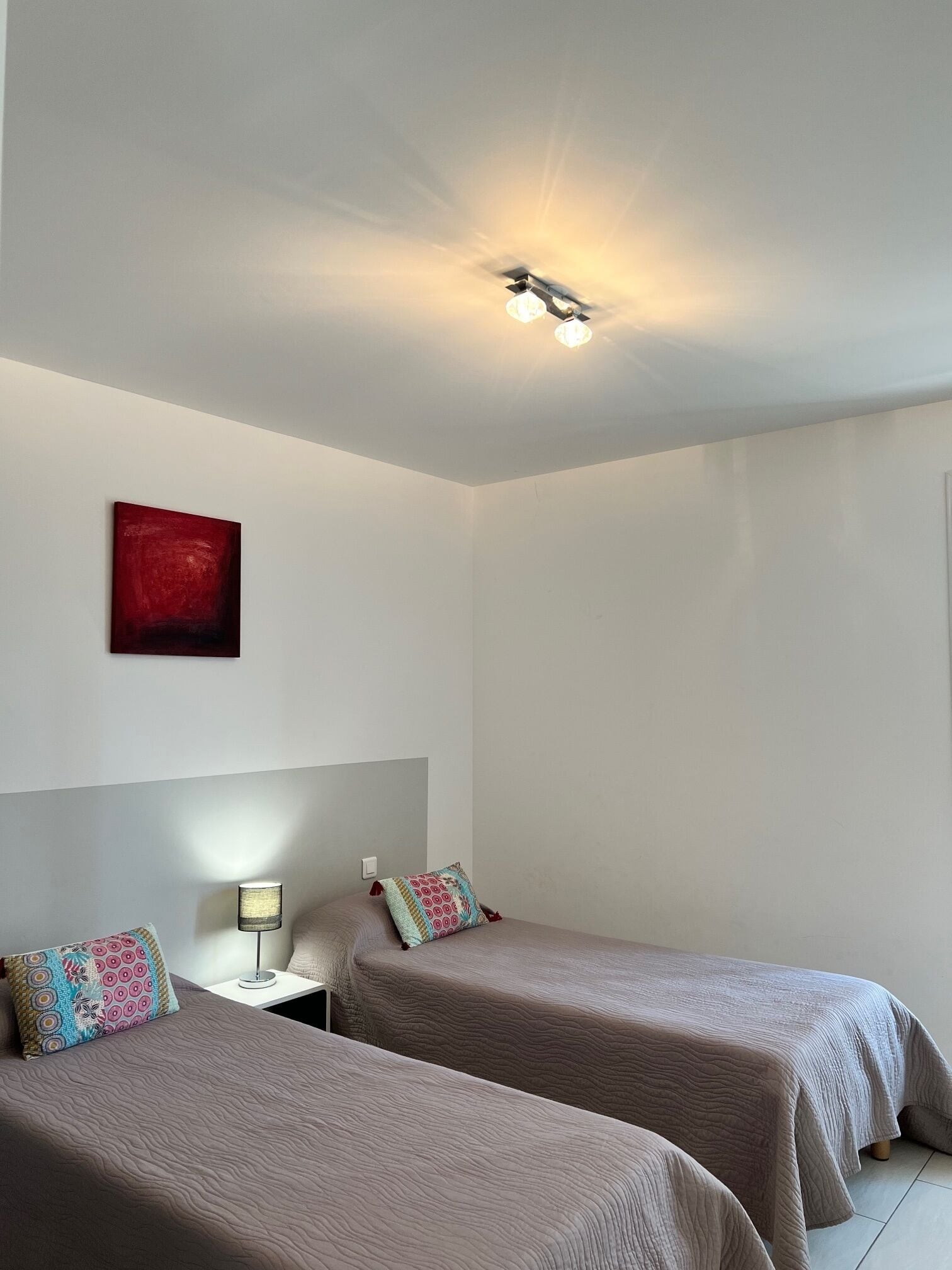 A room with two beds, a nightstand, a lamp, a red abstract painting, and a ceiling light fixture.