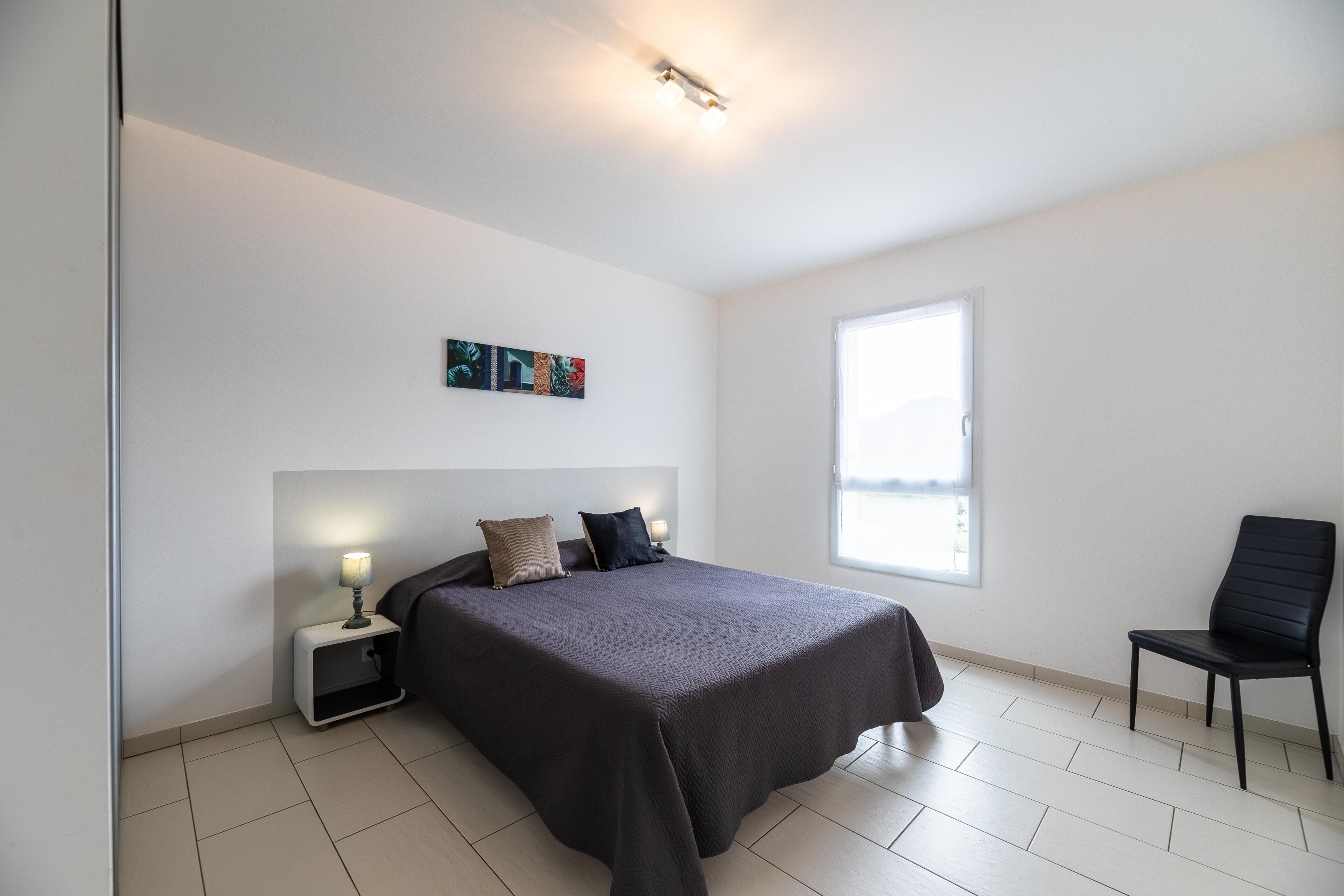 A bedroom with a bed, bedside table, lamp, chair, and a window.