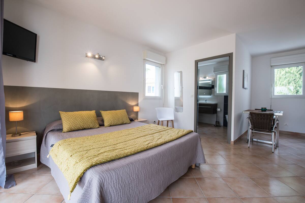 A bedroom with a bed, bedside tables, a TV, and a view into the kitchen.