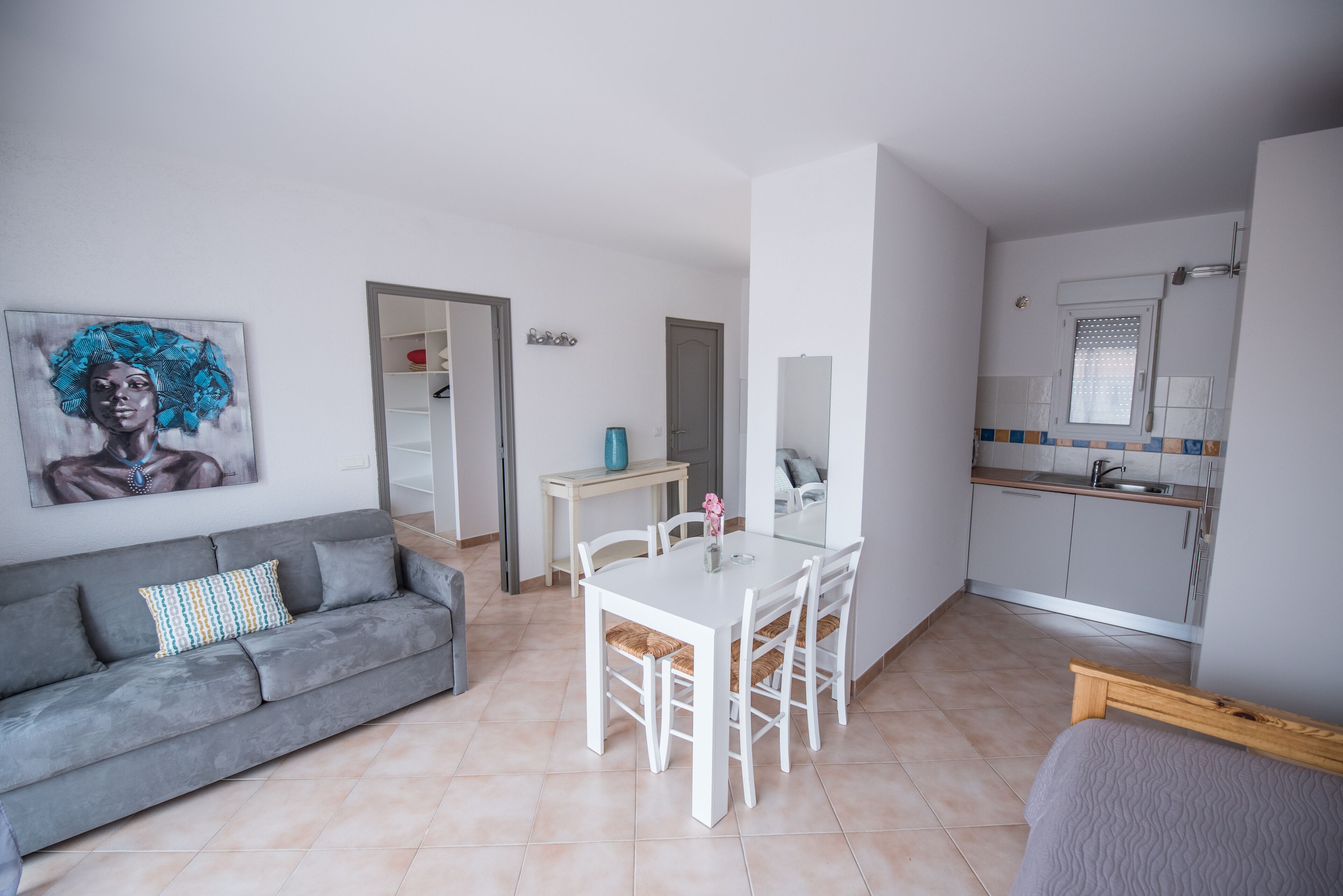 A modern living room with a grey sofa, a white dining table with four chairs, a kitchen area with a sink and cabinets, and a painting on the wall.