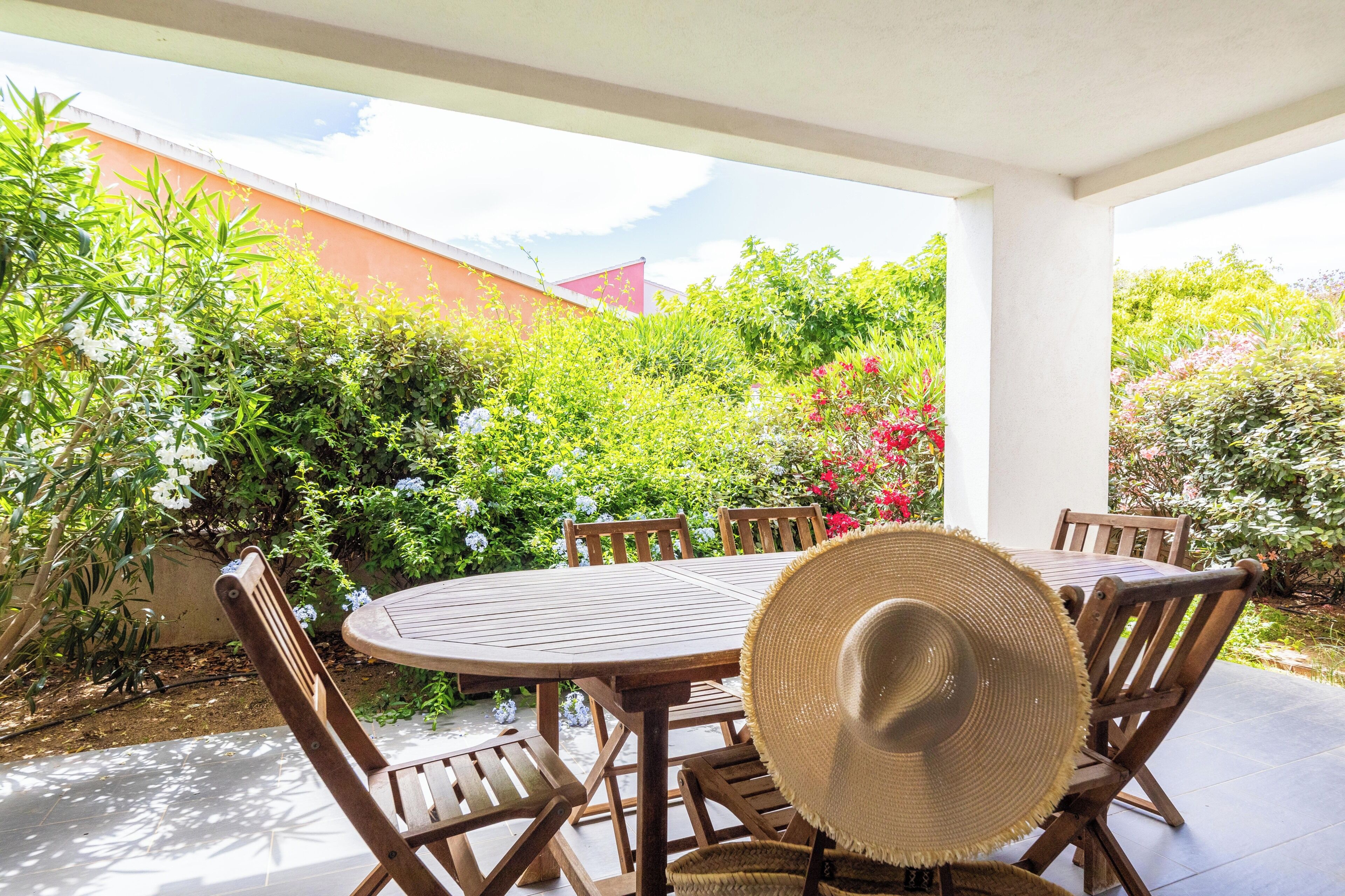 Um pátio externo com mesa e cadeiras de madeira redonda, rodeado por vegetação exuberante e plantas floridas.