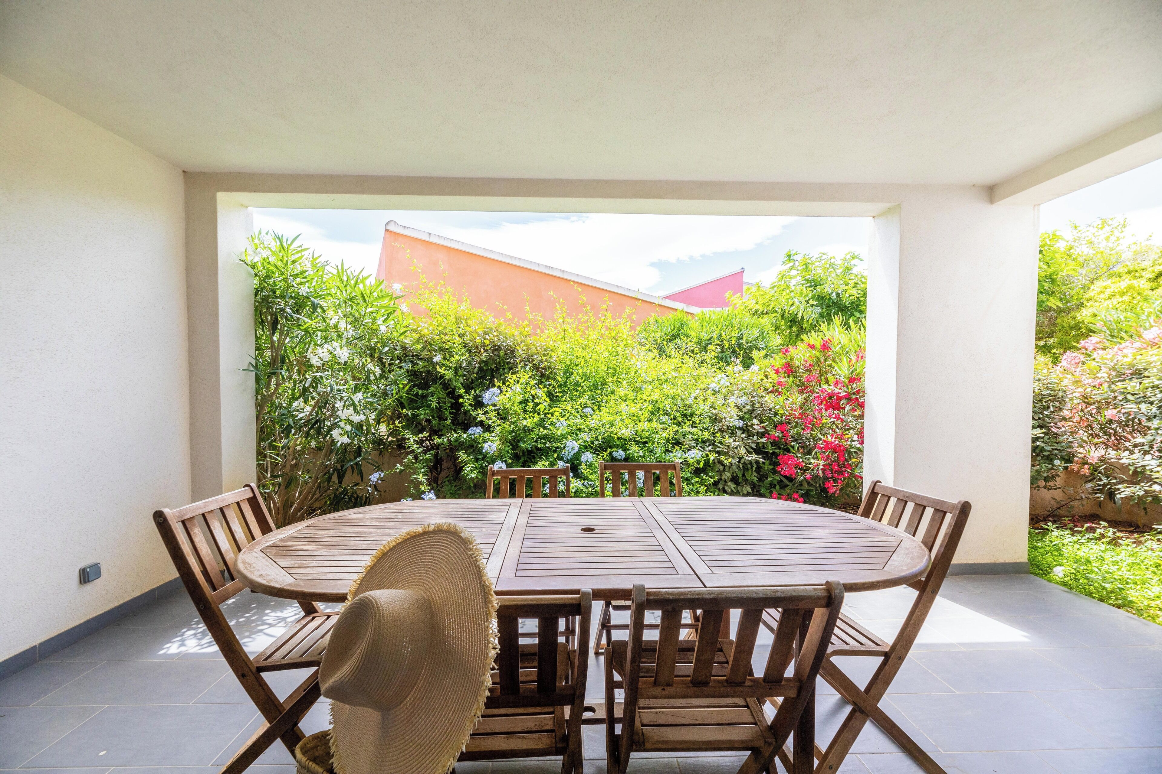 Une table ronde en bois, entourée de chaises, se trouve dans un espace extérieur abrité, d’où l’on peut admirer un jardin et un bâtiment en arrière-plan.