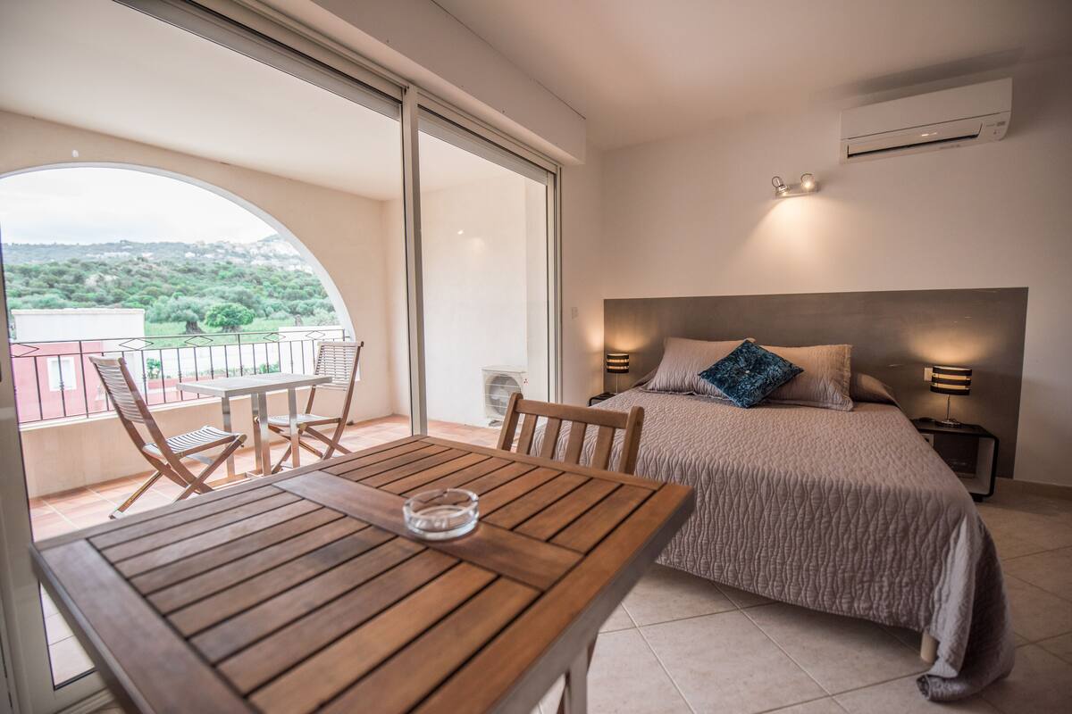A bedroom with a bed, bedside lamps, a table with chairs, and a view through a large window.