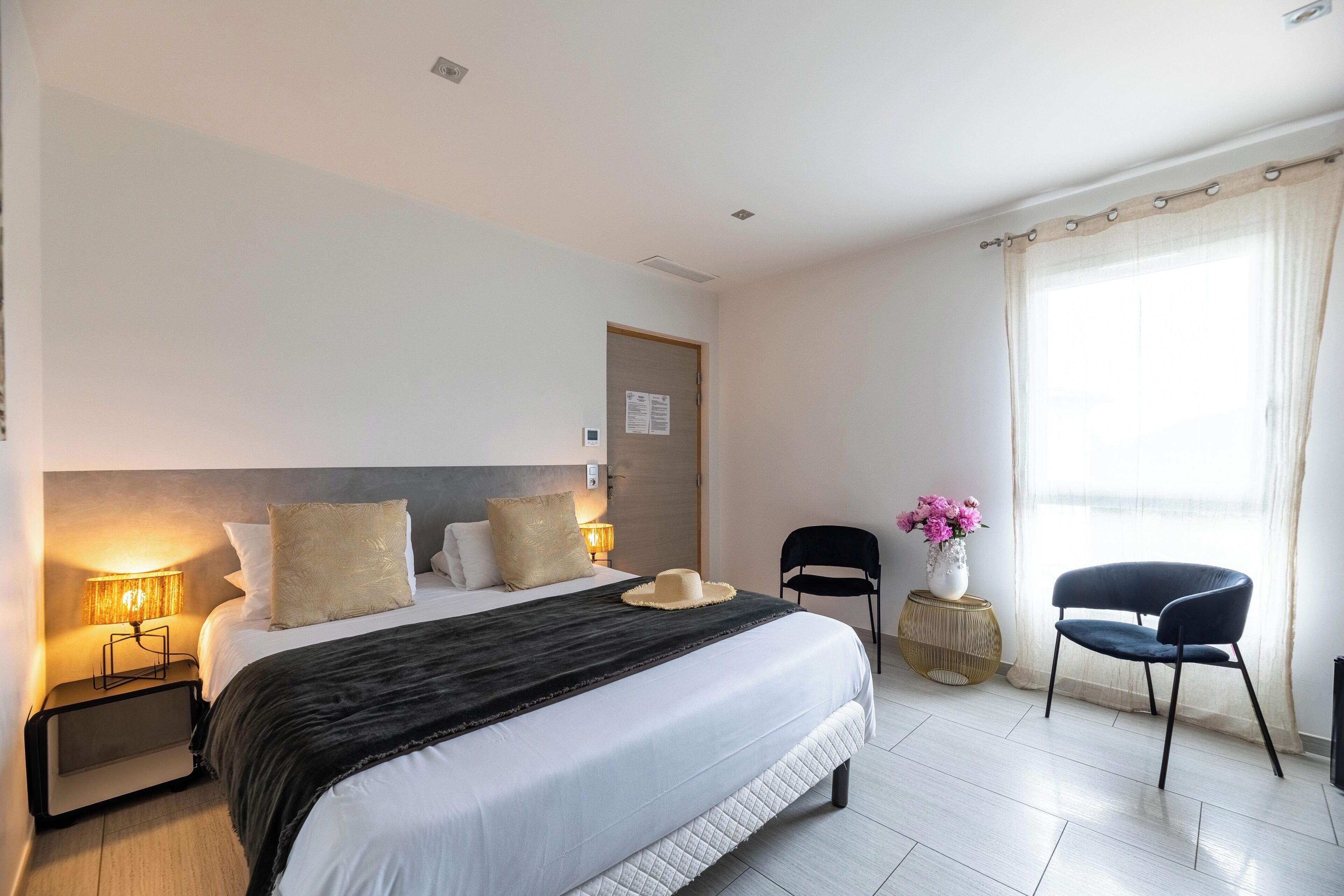 A hotel room with a large bed, bedside lamps, a chair, a small table with flowers, and a window with curtains.