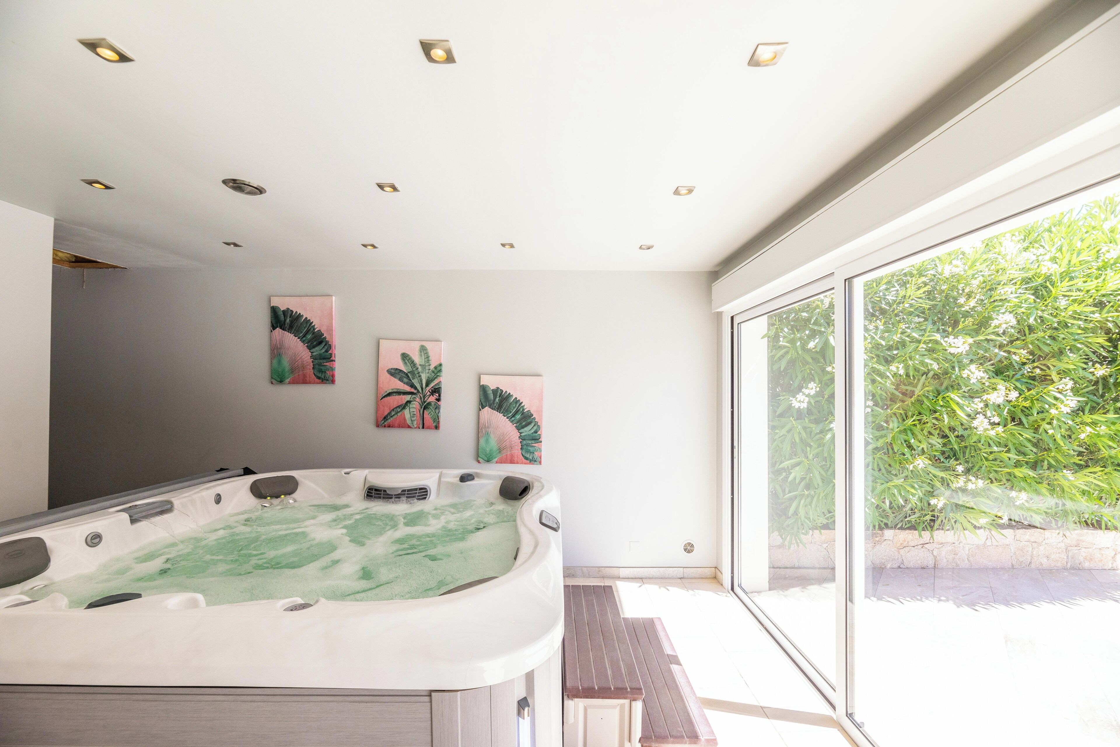 Una bañera de hidromasaje en una habitación con un ventanal grande, luces empotradas en el techo y decoración en las paredes.