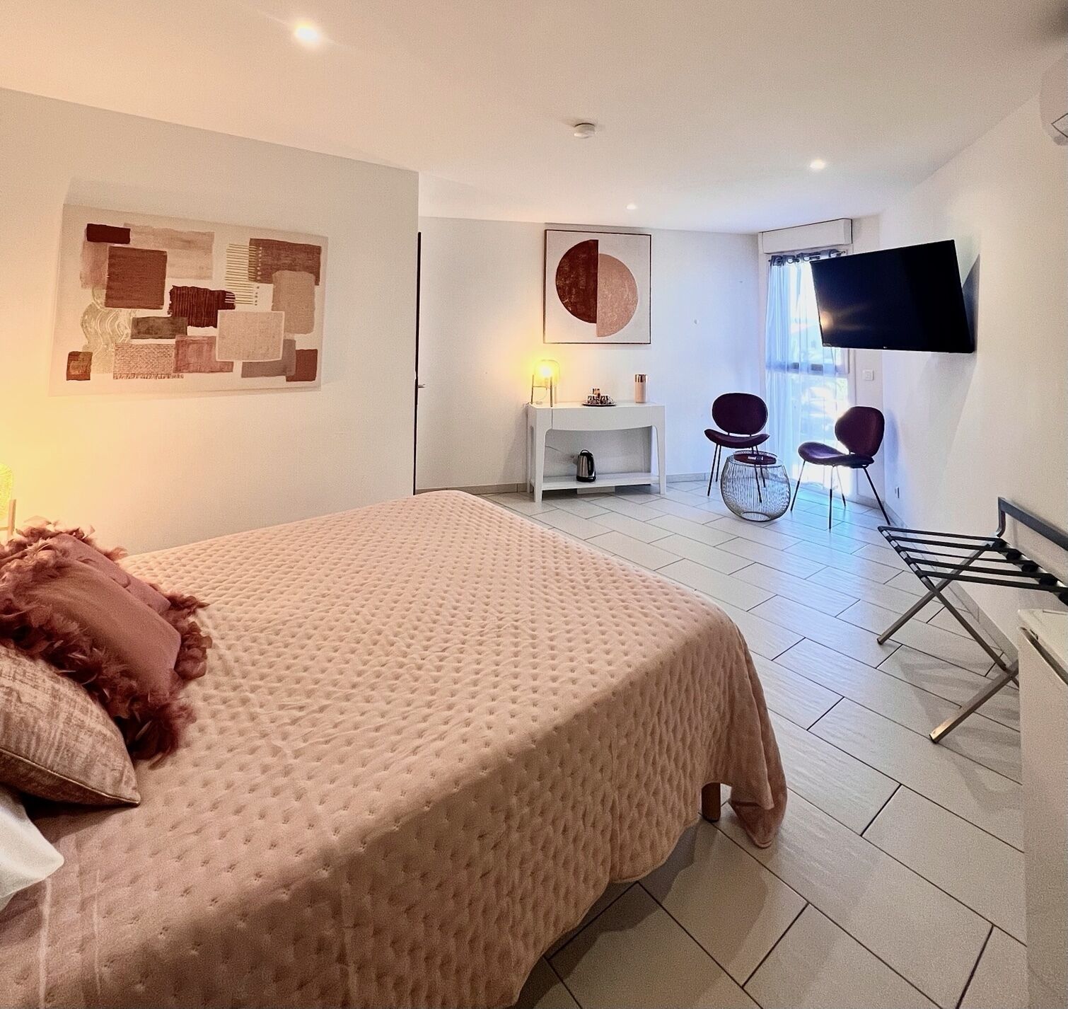 A modern hotel room with a large bed, a flat-screen TV mounted on the wall, a small seating area with chairs, and a window providing natural light.