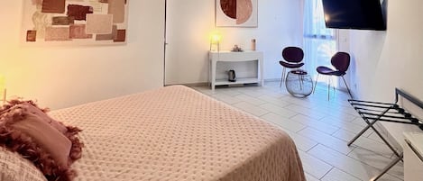 Una habitación de hotel moderna con una cama grande, un televisor de pantalla plana montado en la pared, un pequeño rincón de descanso con sillas y una ventana que deja entrar luz natural.