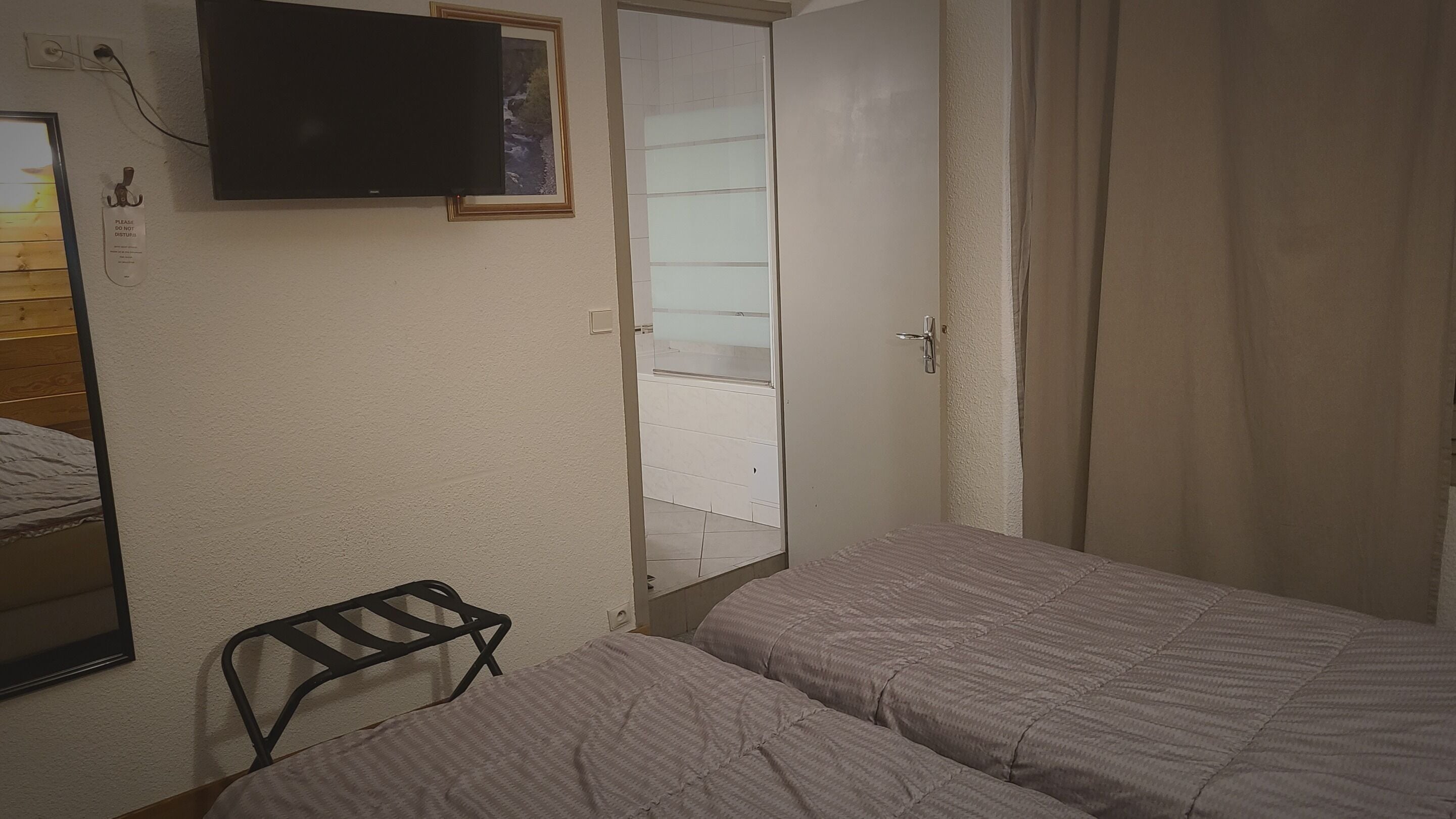 A bedroom with two beds, a TV mounted on the wall, a small chair, and a wardrobe with a mirror.