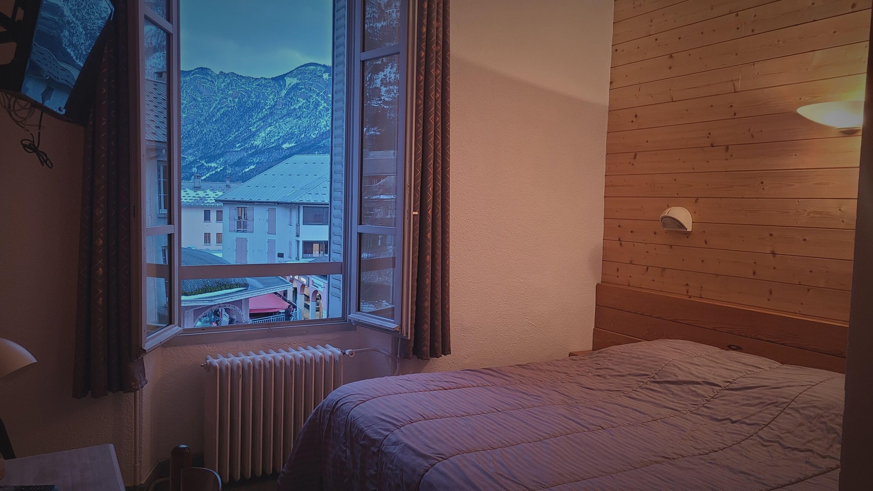 A bedroom with a wooden headboard, a window with a view of buildings and mountains, and a radiator below the window.