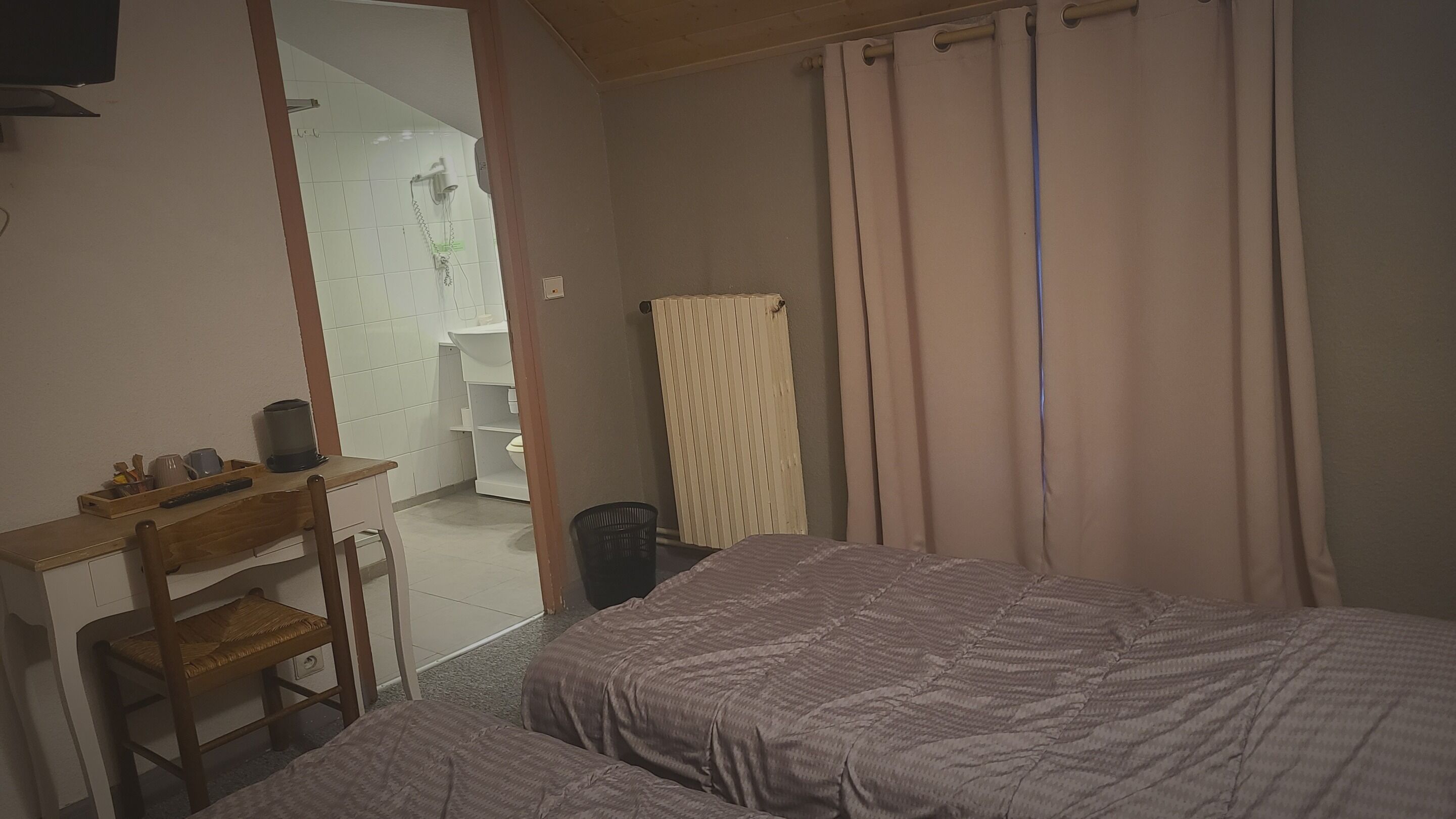 Un dormitorio con dos camas, un escritorio con silla, un radiador y un baño visible a través de una puerta abierta.