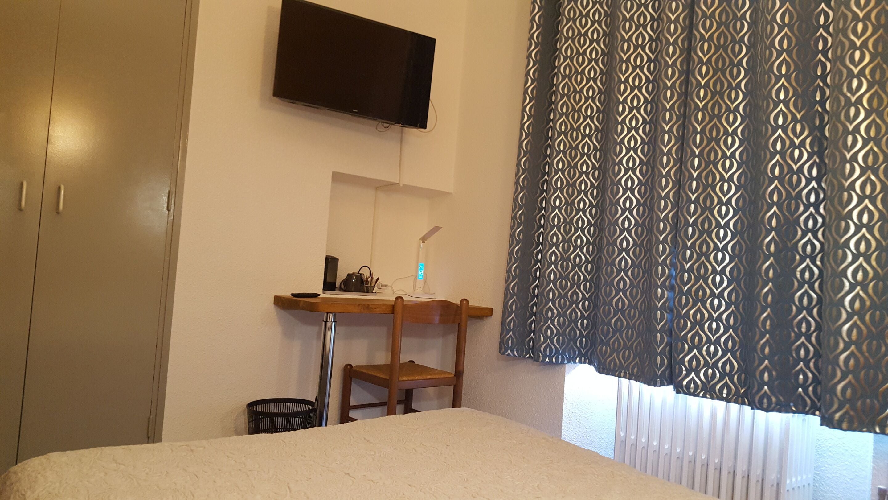 Ein Hotelzimmer mit einem Bett, einem Schreibtisch mit Stuhl, einem an der Wand befestigten Fernseher und gemusterten Vorhängen.