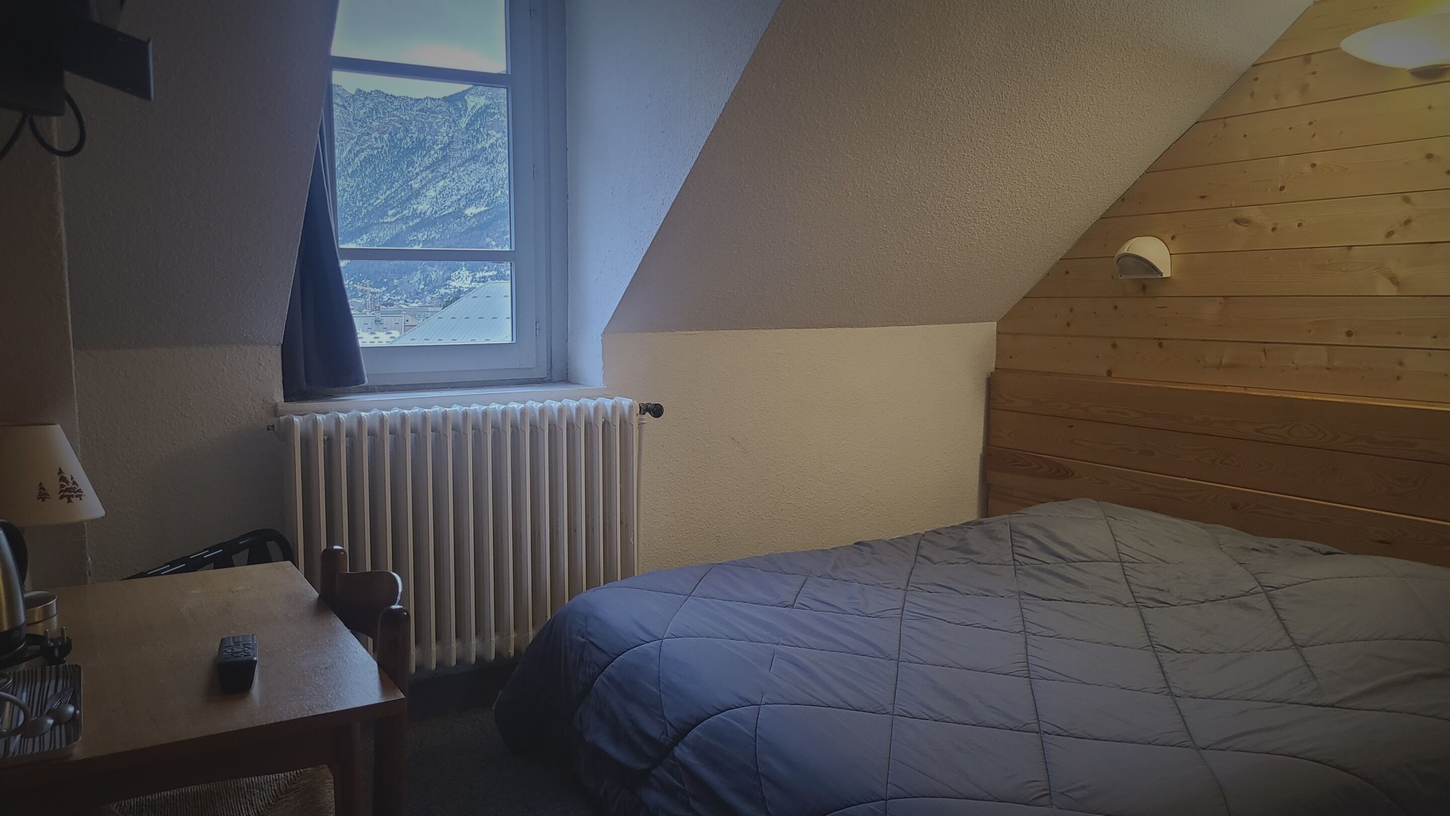 A small, cozy room with a sloped ceiling, a wooden headboard, a bed with a quilt, a desk with a lamp, and a window with a view of mountains.
