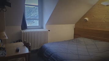 A small, cozy room with a sloped ceiling, a wooden headboard, a bed with a quilt, a desk with a lamp, and a window with a view of mountains.