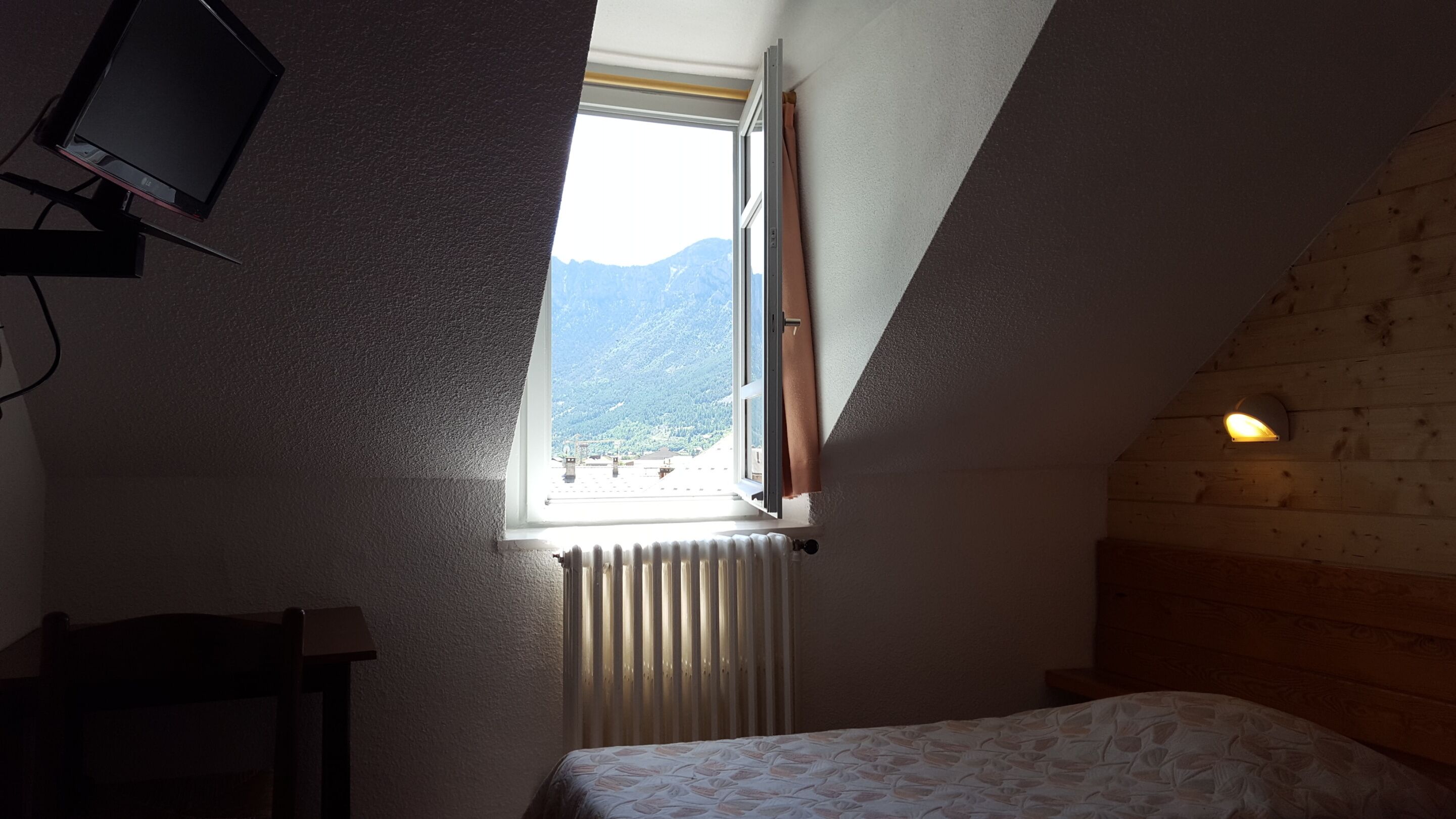 A bedroom with a sloped ceiling, a window with a view of mountains, a bed, a radiator, and a wall-mounted light fixture.