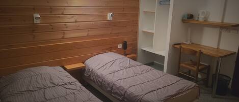 Un dormitorio con dos camas individuales, cabecero de madera, mesita de noche, estante con lámpara y escritorio con silla.