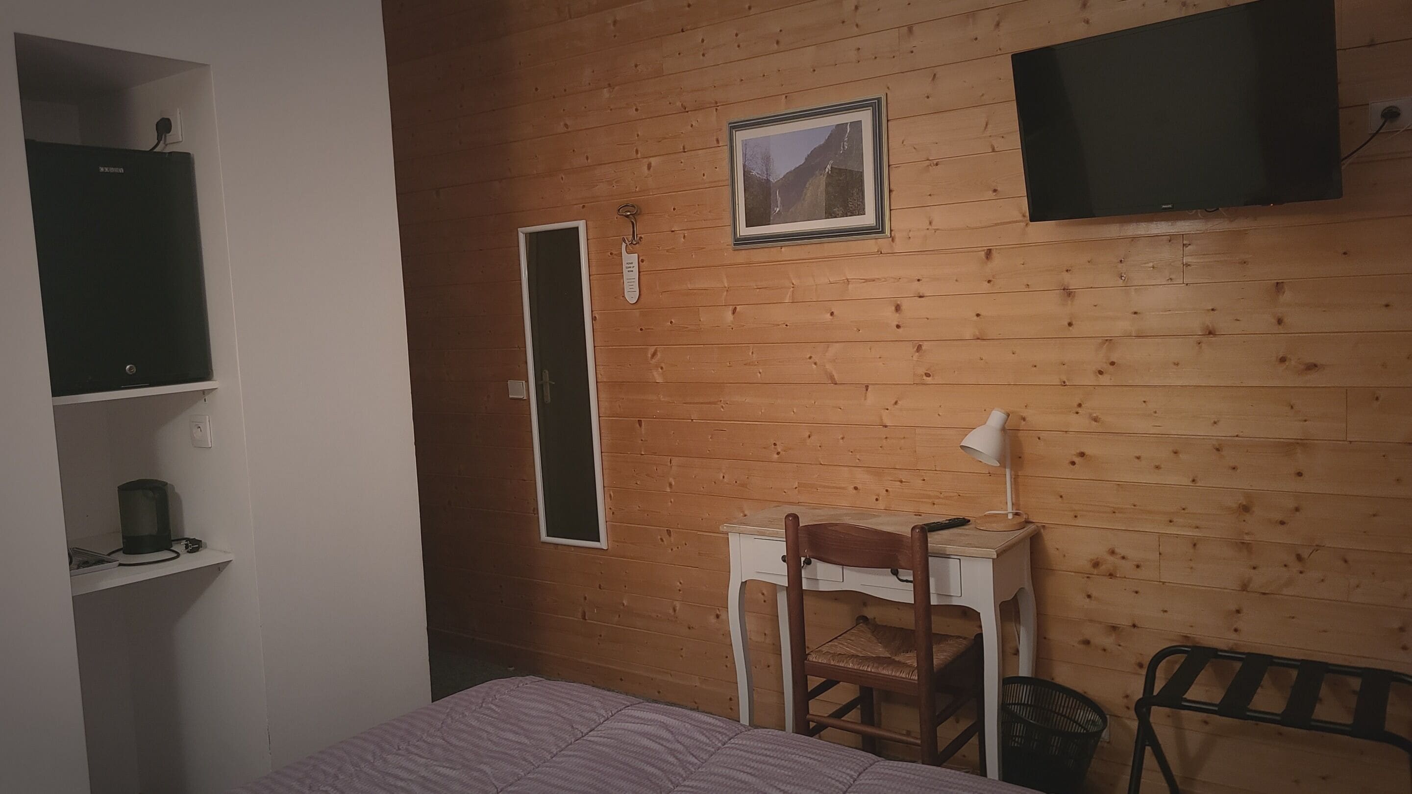 Quarto com paredes de madeira, uma cama, uma escrivaninha com cadeira, uma TV fixada na parede e uma prateleira com chaleira.