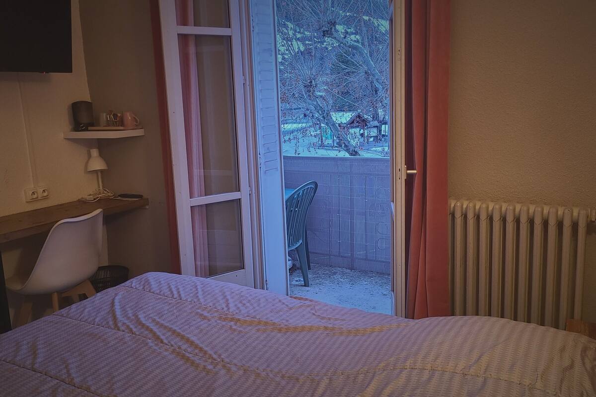 A bedroom with a bed, a desk, a chair, a radiator, and a sliding glass door leading to a balcony with a view of trees and a fence.