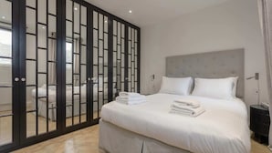 A hotel room with a bed, a nightstand, a large window, and a sliding door with a view of the outside. - Elegant 1 Bedroom Apartment in South Kensington (London)