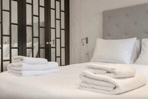 A neatly made bed with white linens and folded towels, a grey upholstered headboard, and a modern, minimalist room design with a frosted glass partition. - Elegant 1 Bedroom Apartment in South Kensington (London)