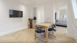 A modern living room with a wooden dining table, two bar stools, a flat-screen TV mounted on the wall, and a sofa in the background. - Elegant 1 Bedroom Apartment in South Kensington (London)
