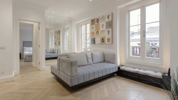 A spacious room with a large grey sofa, a bench, and a wall-mounted artwork. - Elegant 1 Bedroom Apartment in South Kensington (London)