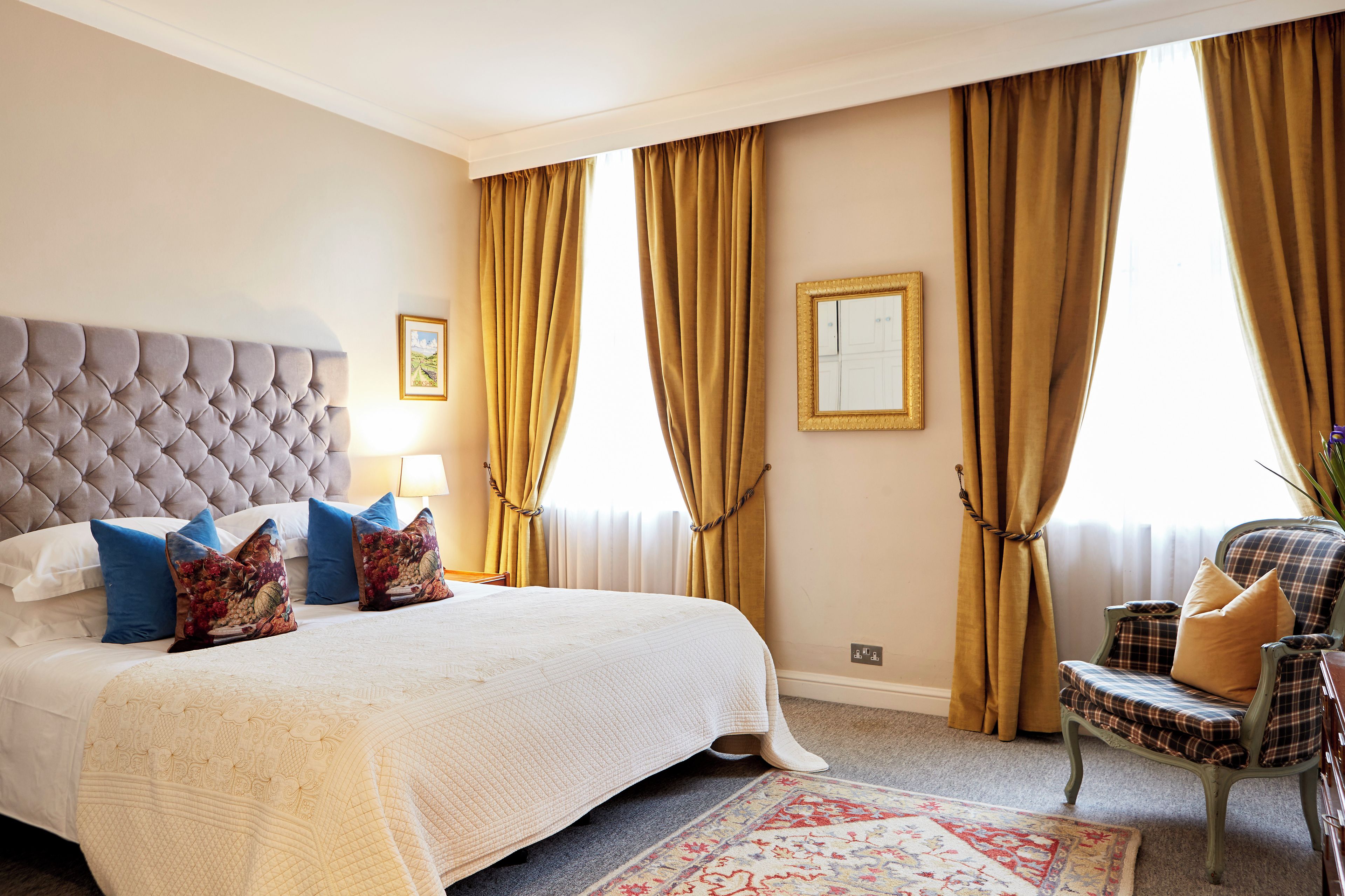 A bedroom with a tufted headboard, a large bed with white bedding and decorative pillows, a chair with plaid upholstery, and a patterned rug.