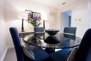 Un comedor con una mesa de vidrio, dos sillas, un florero con flores y dos lámparas fijadas a la pared.