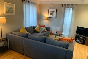 Una sala de estar moderna con un sofá seccional gris, una mesita redonda pequeña, una lámpara de pie, un televisor y dos cuadros enmarcados en la pared.