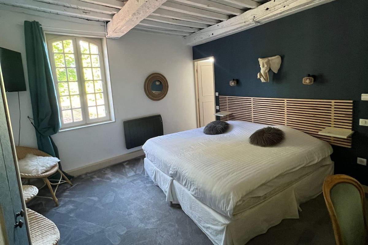 A bedroom with a large bed, a window with curtains, a chair, and a wall-mounted mirror.
