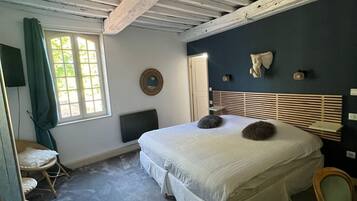 A bedroom with a large bed, a window with curtains, a chair, and a wall-mounted mirror.