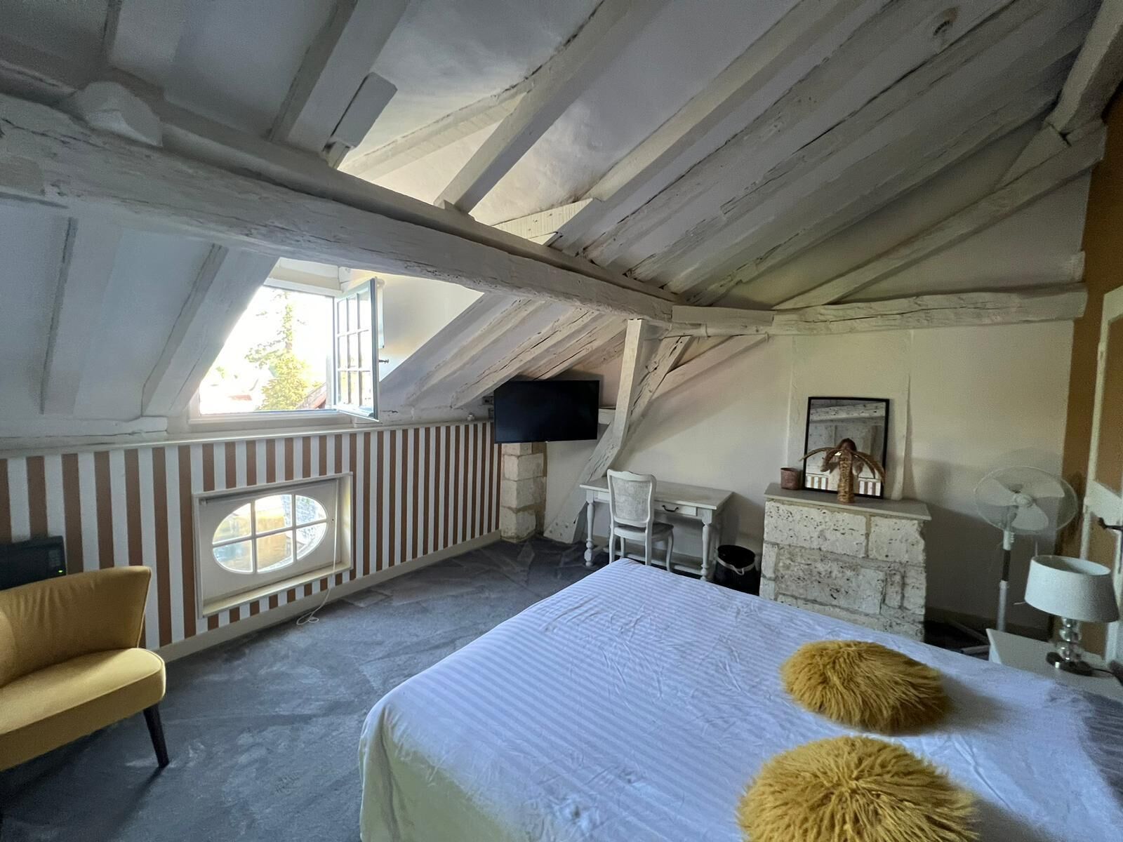 A room with a sloped ceiling, a bed with a white bedspread, two round, fur-like pillows, a small desk with a chair, a television mounted on the wall, and a window with a view of the outside.