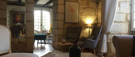 A cozy room with stone walls, a wooden ceiling, and a mix of modern and classic furniture. There are two armchairs, a small table with a glass, and a decorative elephant statue.
