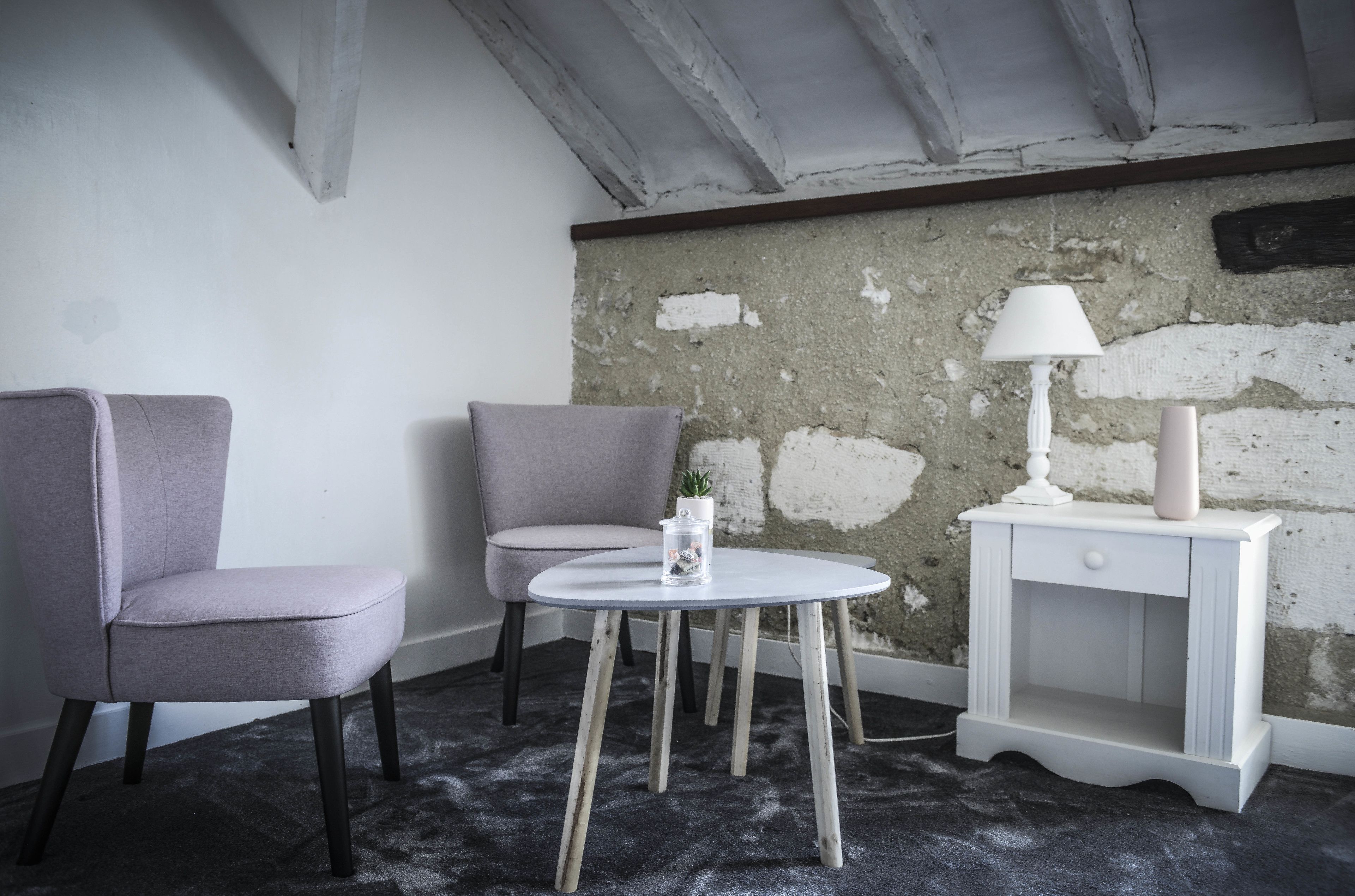 Una habitación con una silla gris, una mesita redonda pequeña con una lámpara y una mesita de noche blanca.