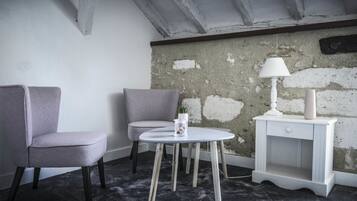 Una habitación con una silla gris, una mesita redonda pequeña con una lámpara y una mesita de noche blanca.