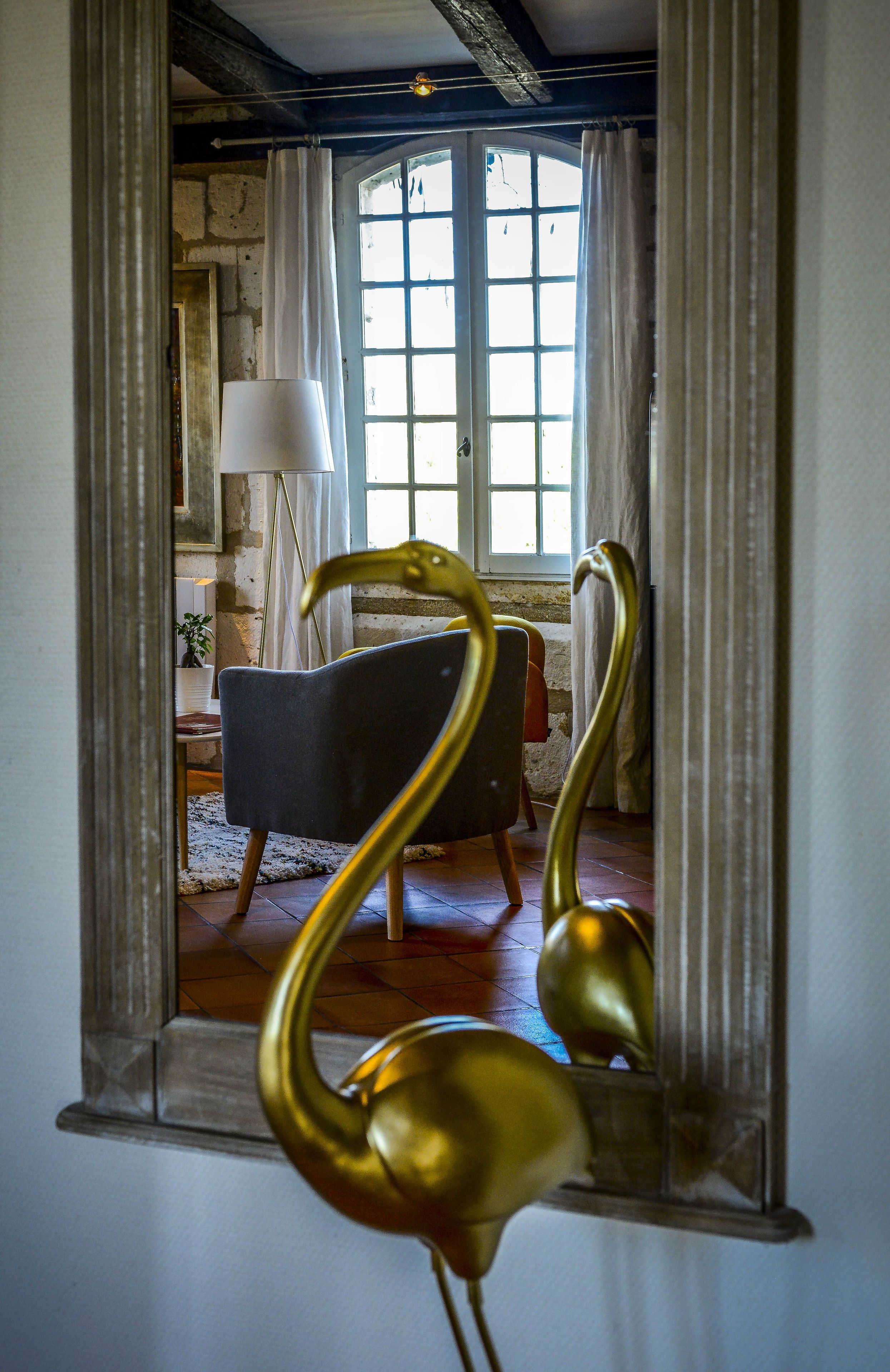 A room with a large mirror reflecting a chair, a lamp, and a window with curtains.