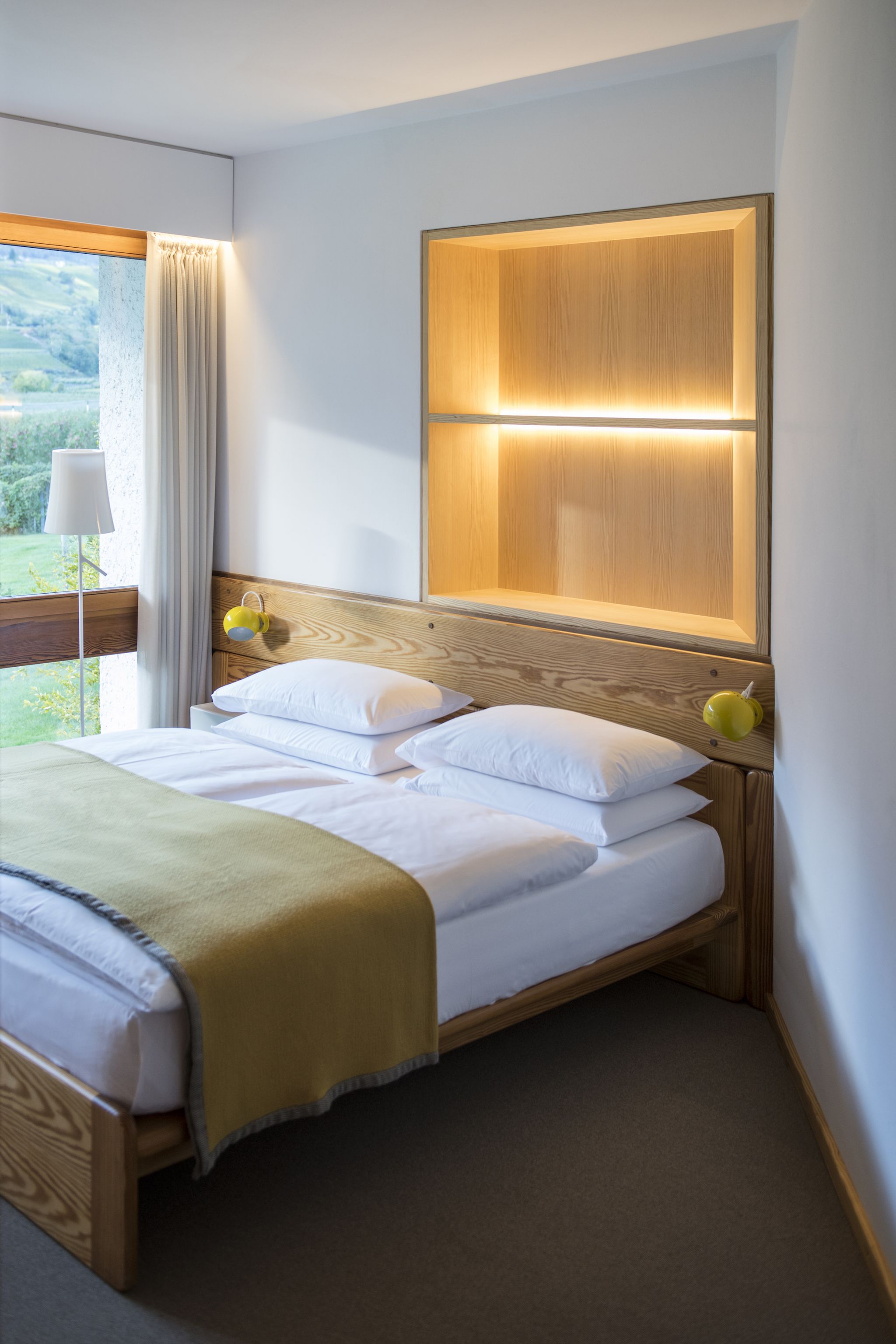 Una cama bien hecha con sábanas blancas y una manta verde, una lámpara de noche y vistas a la vegetación a través de una ventana con cortinas.