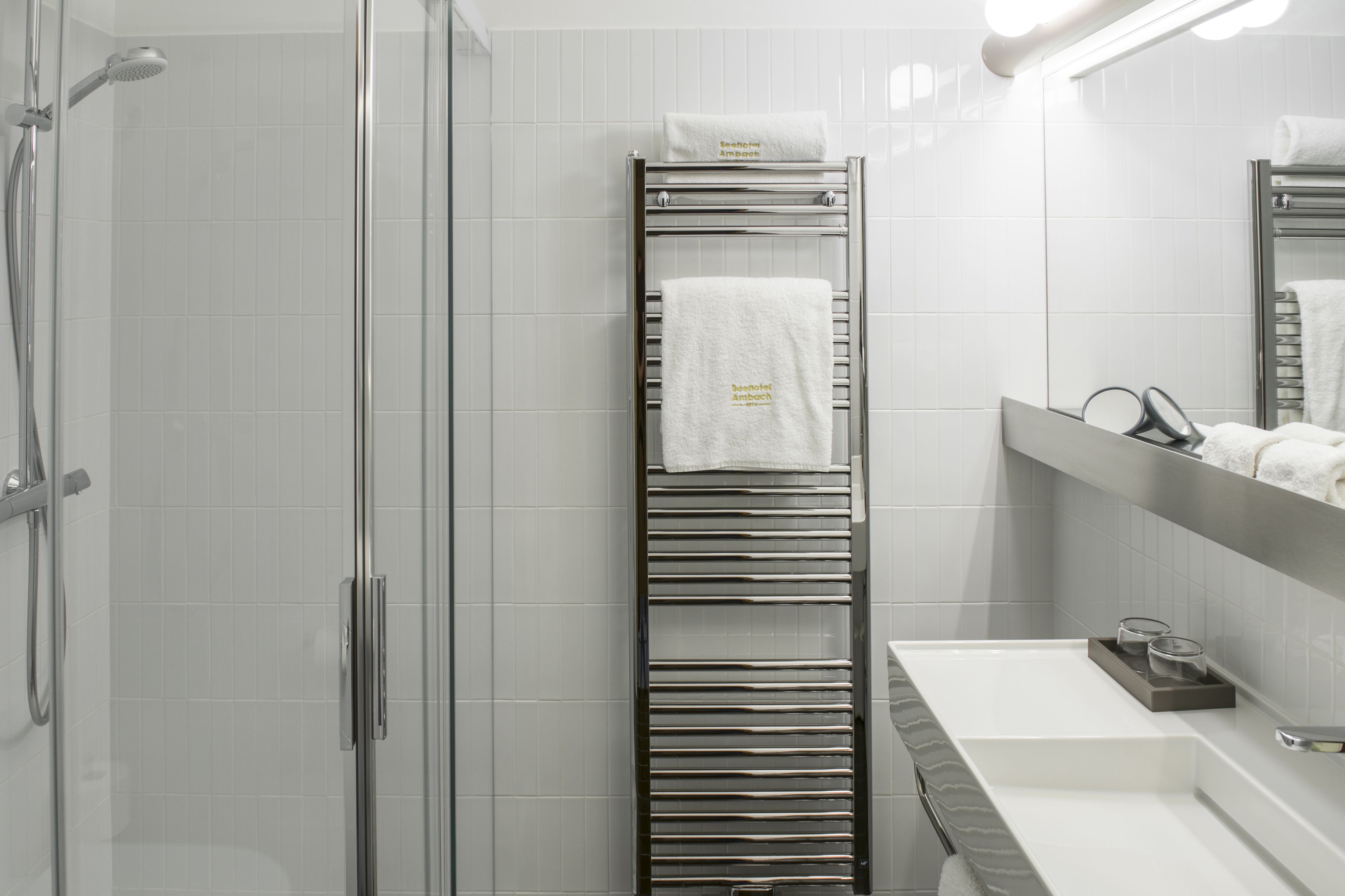 Ein modernes Badezimmer mit Duschkabine aus Glas, einem chromfarbenen Handtuchhalter mit zwei Handtüchern, einem weißen Waschbecken und einem Spiegel darüber.