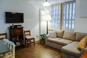 A living room with a flat-screen TV, a bookshelf, a green folding table, a beige sofa, and a window with blinds.