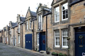 Une rangée de bâtiments traditionnels en pierre, avec des portes et des fenêtres bleues, une rue pavée et de petites plantes en pot à l’entrée.