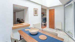 A modern living room with a dining table, a large bowl, and a blue mat. There is a television in the background and a wooden door.