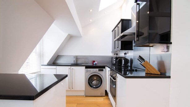A modern kitchen with a washing machine, microwave, and black countertops. - Contemporary 1 Bedroom Flat in Fulham near The Thames (London)