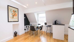A modern kitchen with a central island, a dining area with a glass table and chairs, and a staircase leading to an upper level. - Contemporary 1 Bedroom Flat in Fulham near The Thames (London)