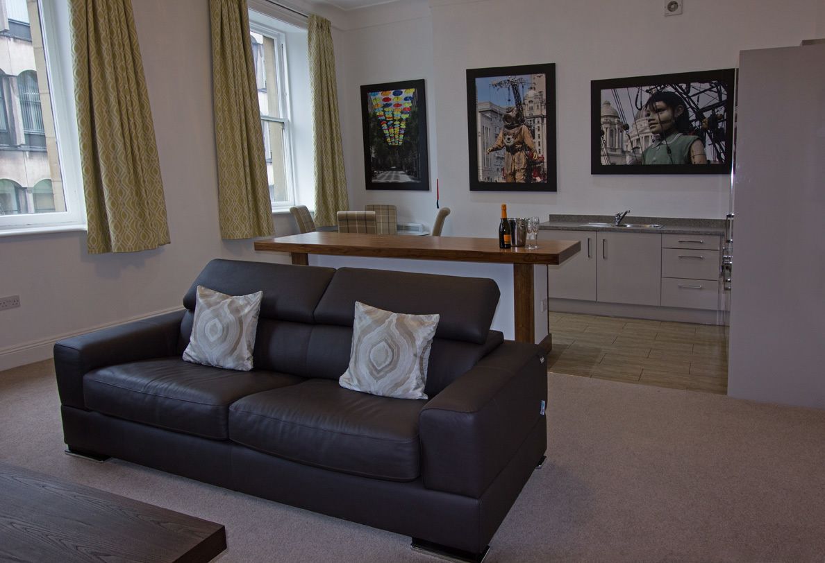A modern living room with a dark leather sofa, a wooden dining table, and a kitchen area with white cabinets.
