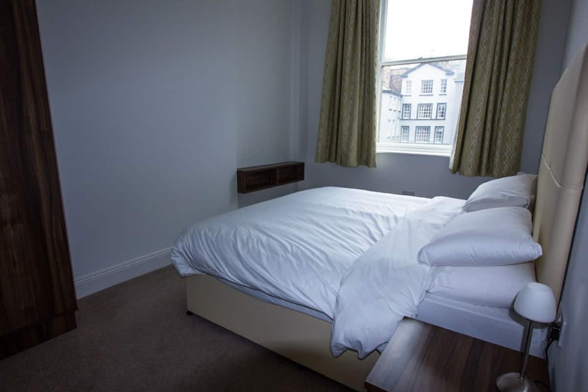 A bedroom with a bed, a bedside table, a window with curtains, and a view of buildings outside.
