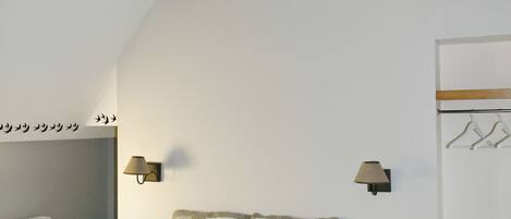 A bedroom with a bed, wooden beams, a wall-mounted light fixture, a radiator, a nightstand, a bench, and a coat rack.