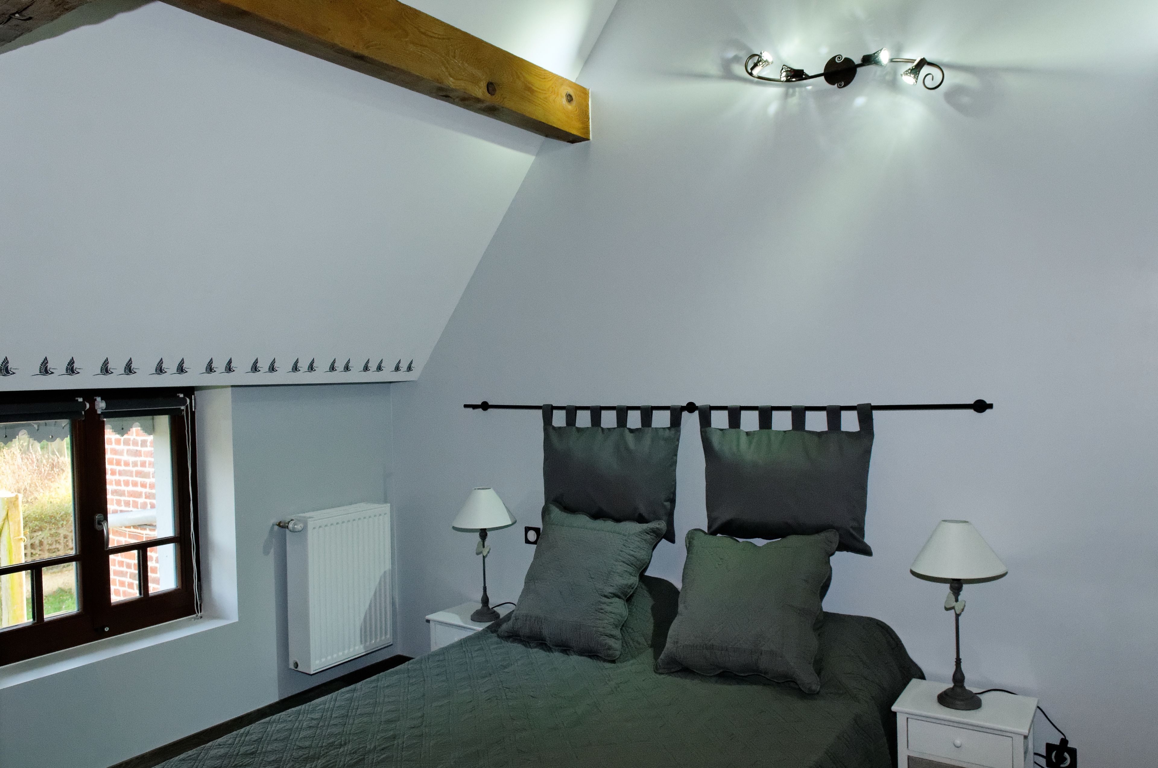 A bedroom with a bed, two bedside tables with lamps, a radiator, a window with a view, and a sloped ceiling with exposed beams.