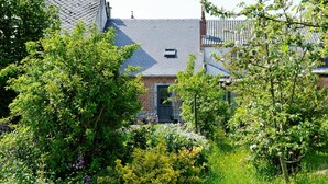 A house with a sloped roof, surrounded by lush greenery and flowering plants. - La Ferme Aux Charmes (Solre-le-Château)